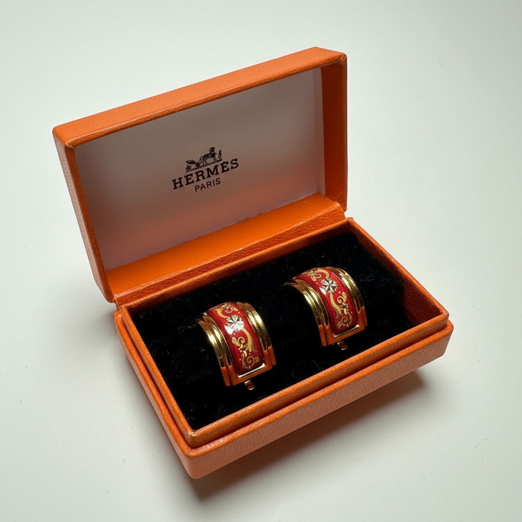 Paire de clips d'oreilles Hermès vintage en émail rouge et or, modèle large, présentés ouverts dans leur boîte orange d'origine.