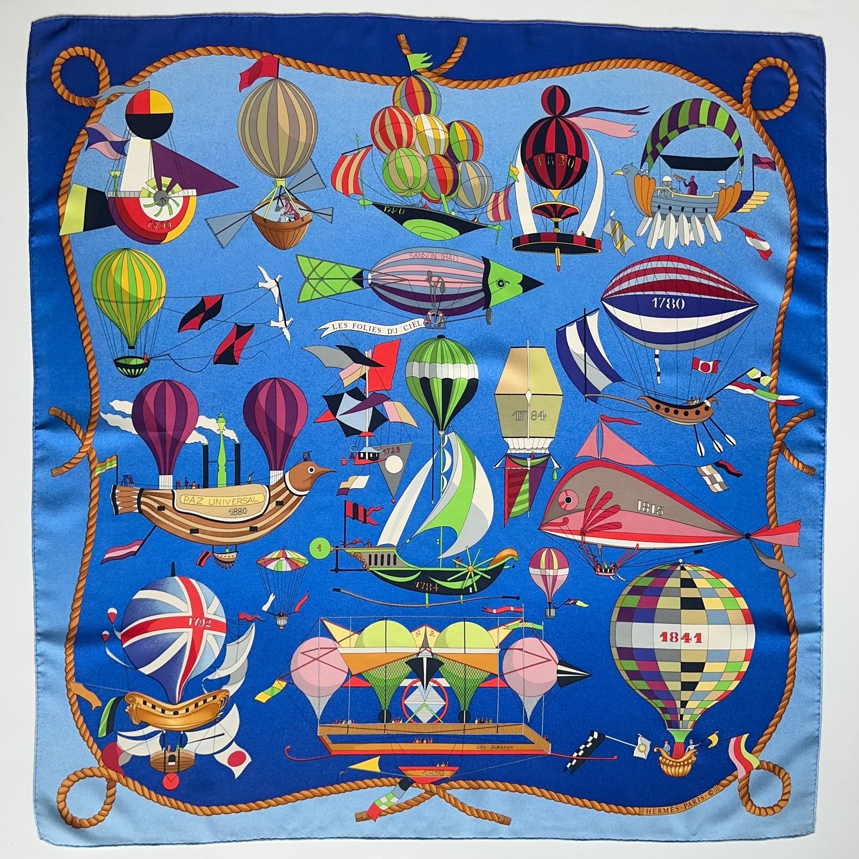 Carré Hermès "Les Folies du Ciel" sur fond bleu roi, illustrant une multitude de montgolfières et machines volantes historiques multicolores.